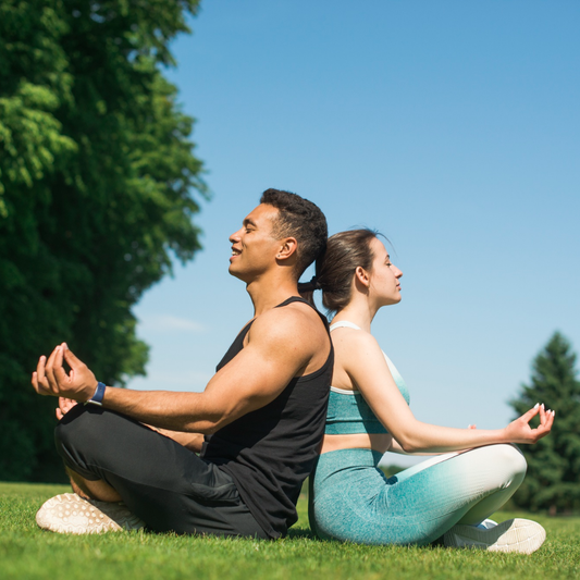 Two people meditating on grass with trees and blue sky in the background