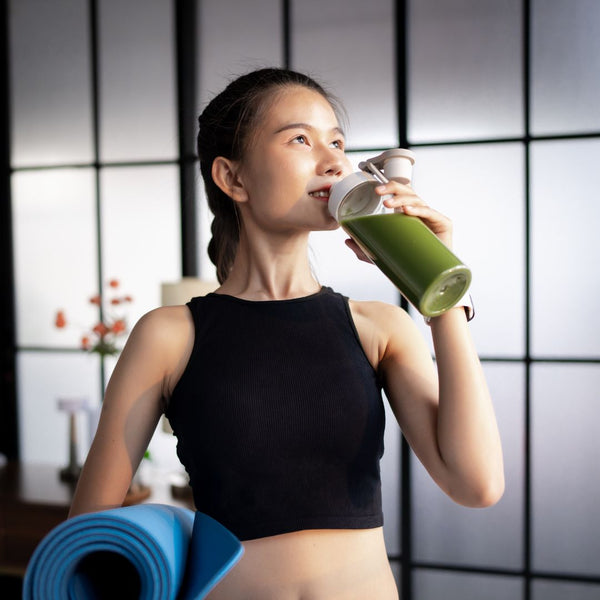 Woman in black athletic top drinking green juice with a blue yoga mat in front of a tiled wall.
